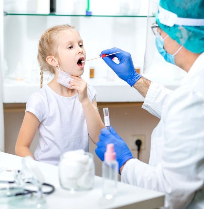 Girl with lowered protective face mask opening mouth for saliva sampling, doctor wearing protective face mask, visor and surgical gloves due to COVID-19 pandemic, PCR DNA testing protocol