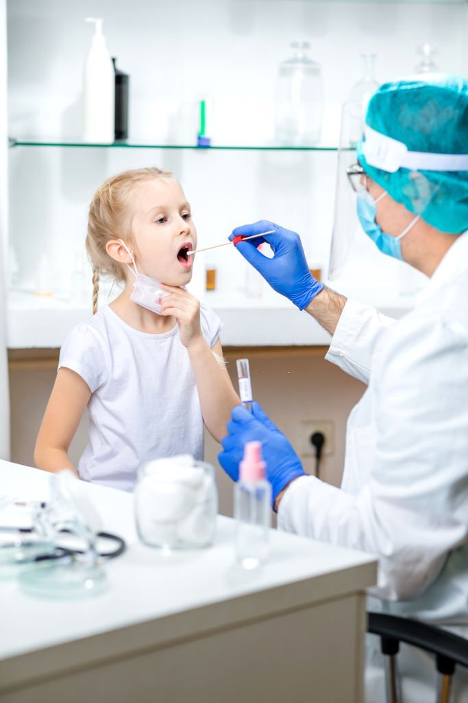 Girl with lowered protective face mask opening mouth for saliva sampling, doctor wearing protective face mask, visor and surgical gloves due to COVID-19 pandemic, PCR DNA testing protocol