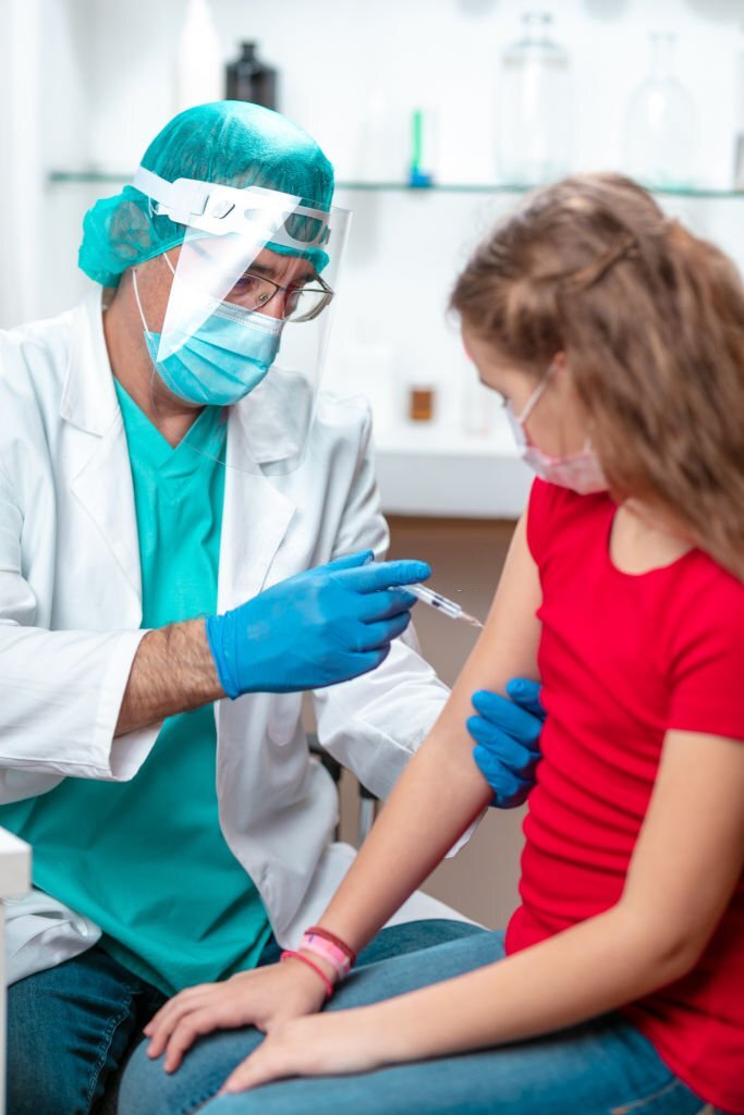 Doctor wearing protective face mask, visor and surgical gloves holding girl's arm with care and injecting COVID-19 vaccine